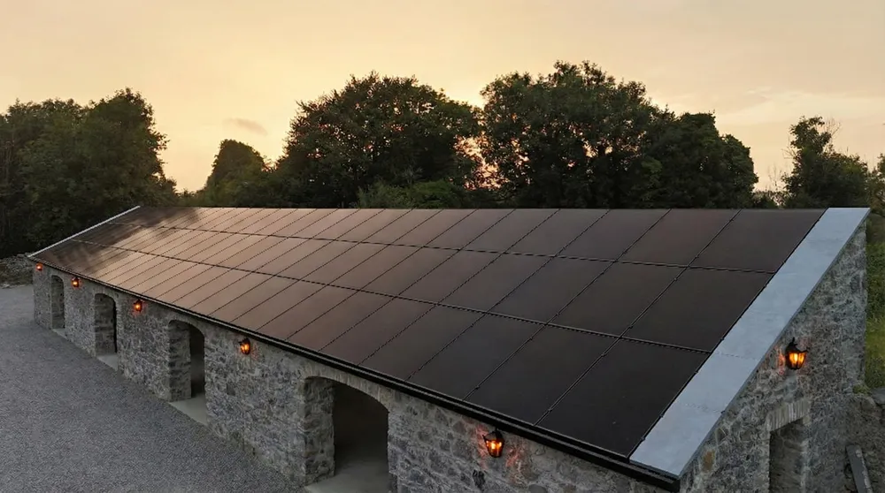 Black in-roof solar panels covering the full pitch of a restored stone outbuilding at sunset, lanterns lit along the wall below